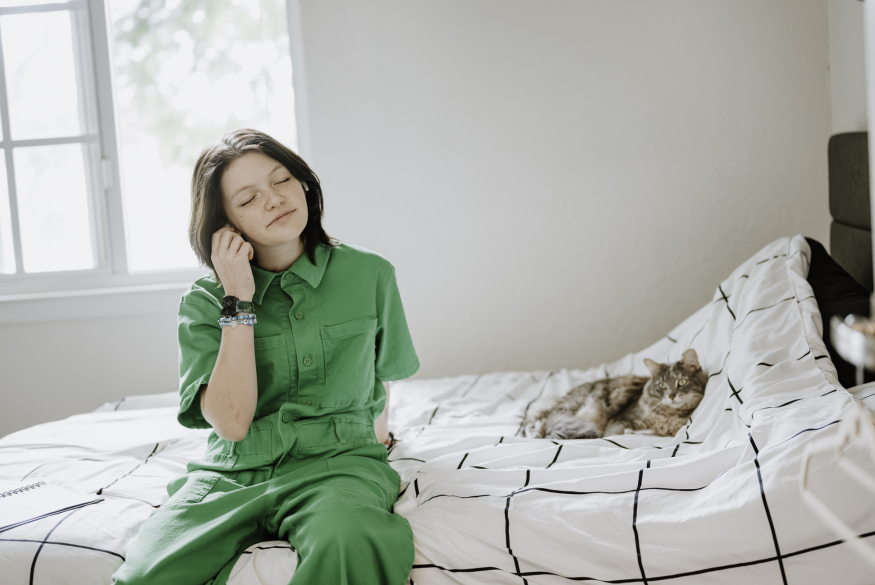 Jongere in groene jumpsuit die naar muziek luistert op bed met kat ernaast