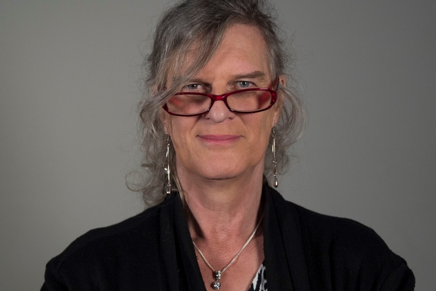 Foto van een vrouw. Ze draagt een rode bril, lange oorbellen en heeft grijze haren die in een staart zijn samengebonden.