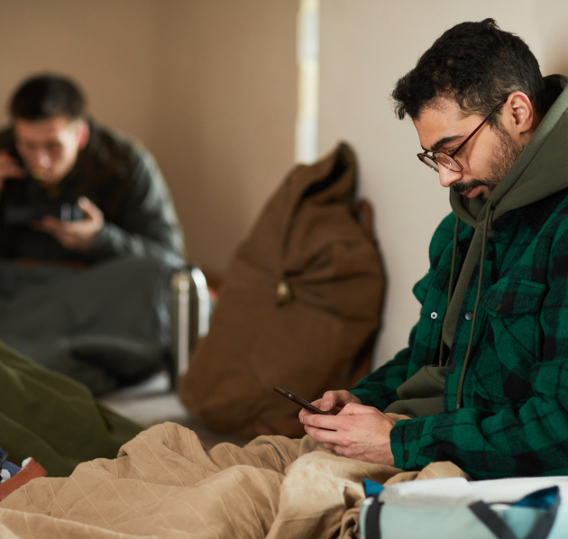 Persoon die neerzit in een asielcentrum en hun gsm vastheeft met links een persoon op de achtergrond die aan het eten is