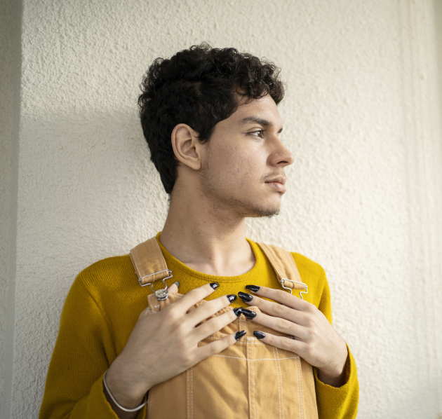 Een jonge persoon met gele salopette, gele trui en zwarte nagellak kijkt naar buiten.