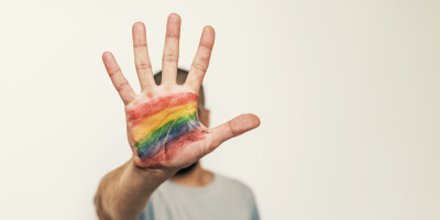 Iemand maakt het stopteken met hun hand. Op het hand staat een regenboogvlag gekleurd. 