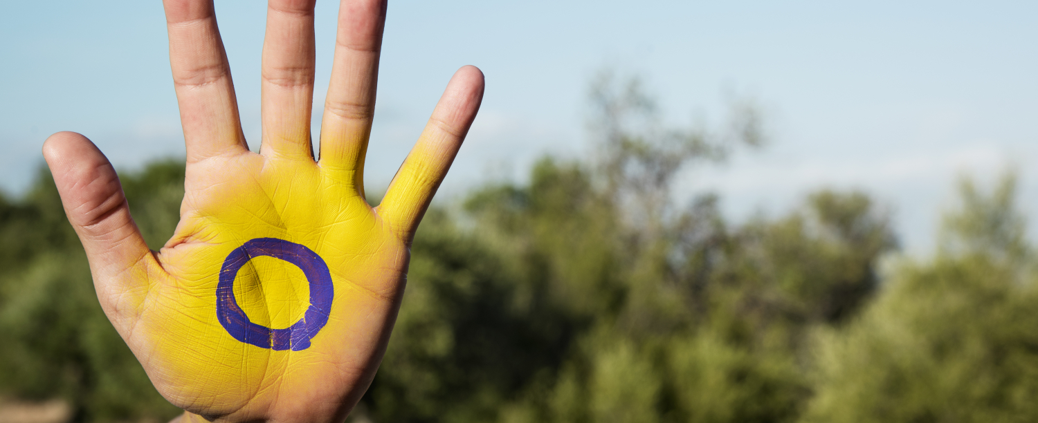 Iemand toont hun hand in de stop houding. Hun hand is geel geschilderd met een paarse cirkel in het midden, het symbool voor de intersekse vlag. 