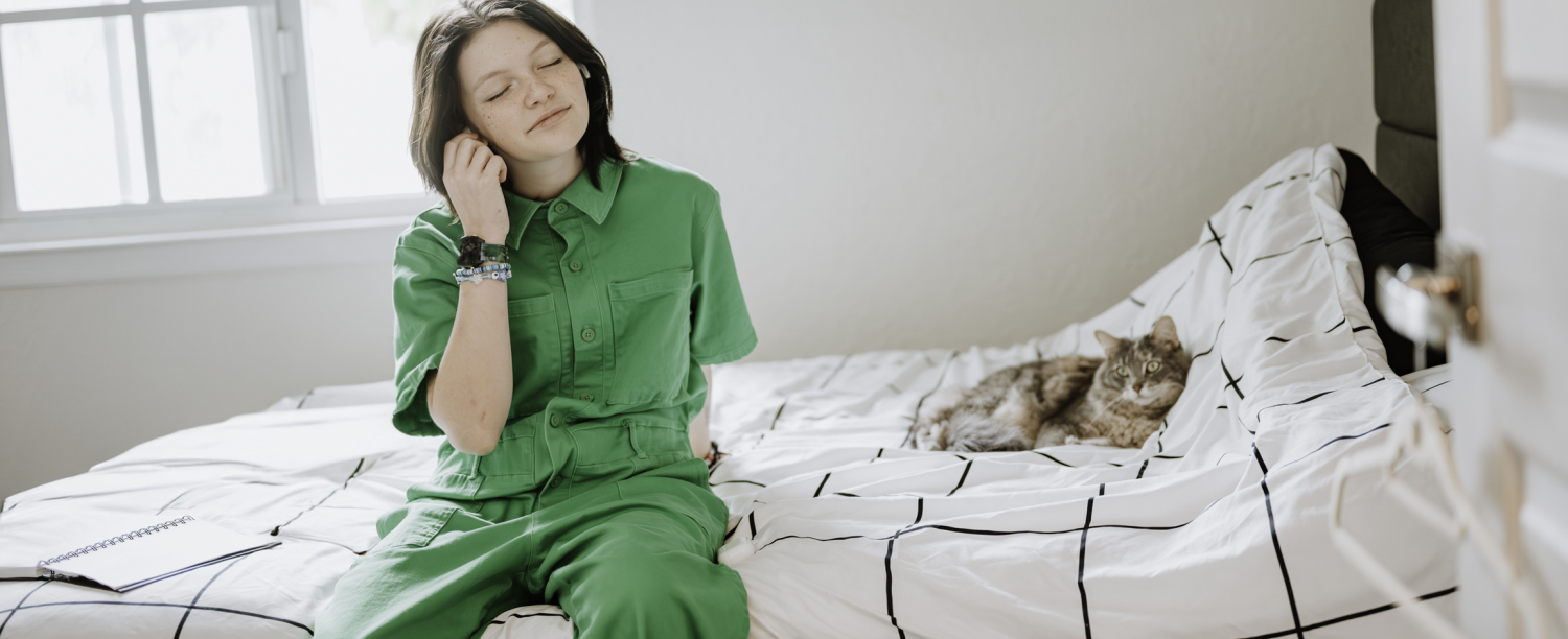 Jongere in groene jumpsuit die naar muziek luistert op bed met kat ernaast